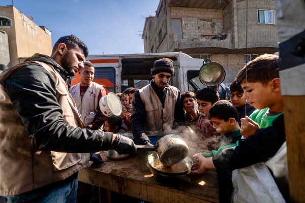 At an outdoor kitchen surrounded by partially destroyed buildings, workers in tan WFP vests ladle up steaming hot pulses into bowls thrust out by hungry children. Photo: WFP/Maxime Le Lijour