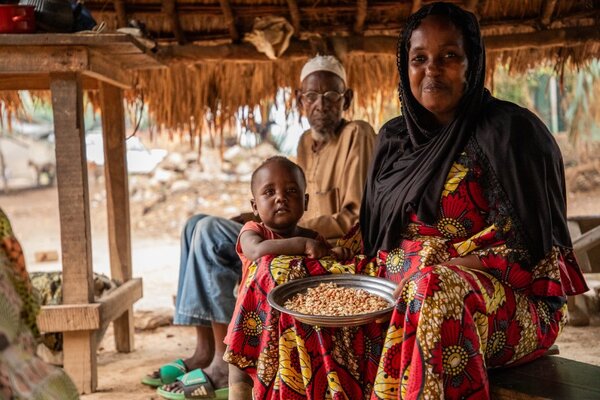A woman in a colourful dress holds a bowl of grains while sitting with a child, with an older man beside them under a thatched shelter in a rural setting.