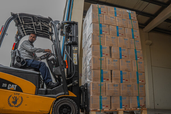 Forklift operator lifts a tall pallet of WFP fortified biscuits wrapped in plastic, stacking boxes inside a warehouse with organised food aid supplies.