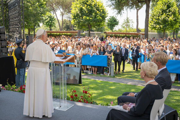 Pope addresses large outdoor crowd from a podium, with attendees gathered behind barriers and trees and greenery surrounding the event space.