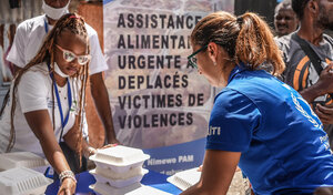 A woman in blue WFP shirt in Haiti helps female colleague across the table box hot meals on a sunny day