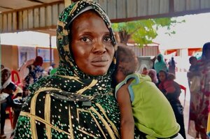 A person wearing a patterned green and black garment holds a small child in a green sleeveless top at a community gathering under a metal-roofed shelter.