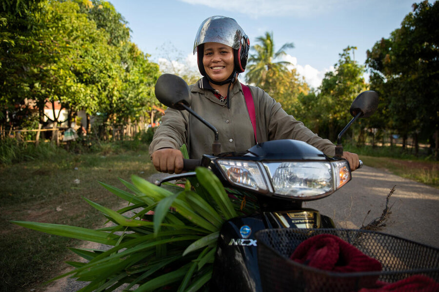 Cambodian woman smiles as she drives a motorbike loaded with green leafy vegetables