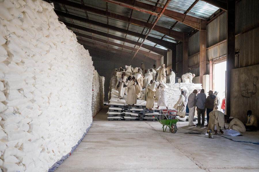 Figures clad in white at the end of a warehouse handle bags of wheat forming a wall to the left of the frame