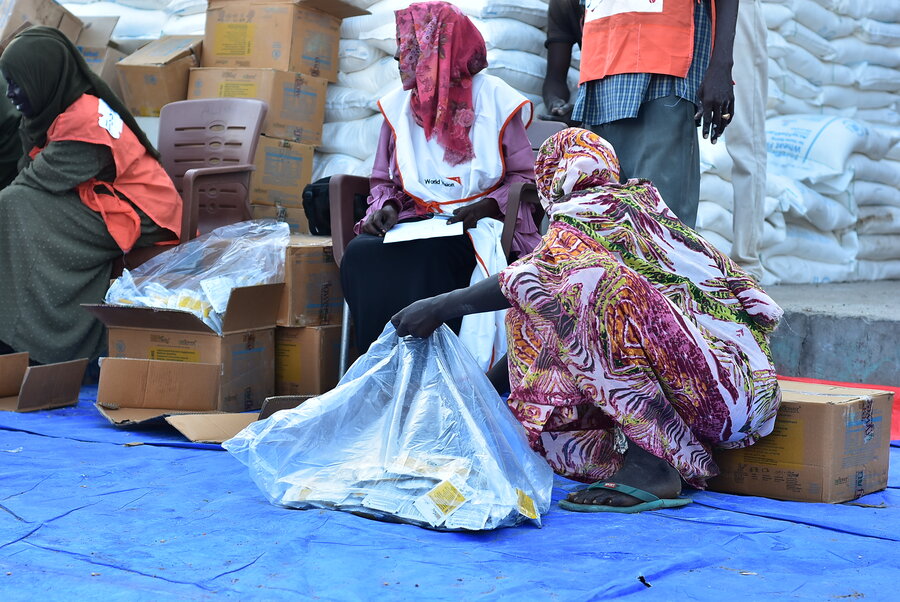 A crouched women in a colourful gown and headscarf holds a plastic bag of WFP food assistance at a distribution point in Kadugli, Sudan. Photo: WFP/Mohamed Elamin