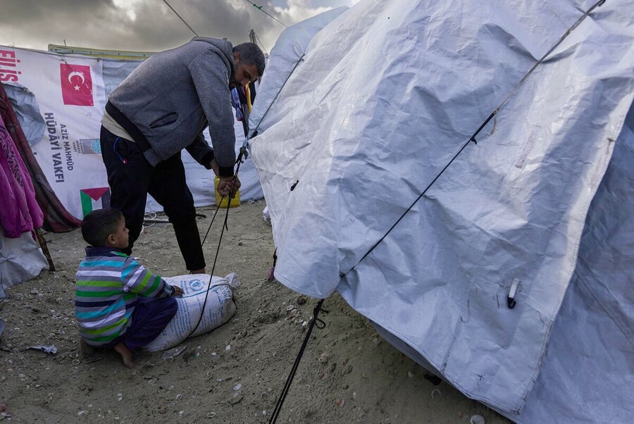 A man and his little boy stand in the sand, surrounded by a tent camp, trying to secure WFP food from winter rains. Photo: WFP/Jaber Badwan