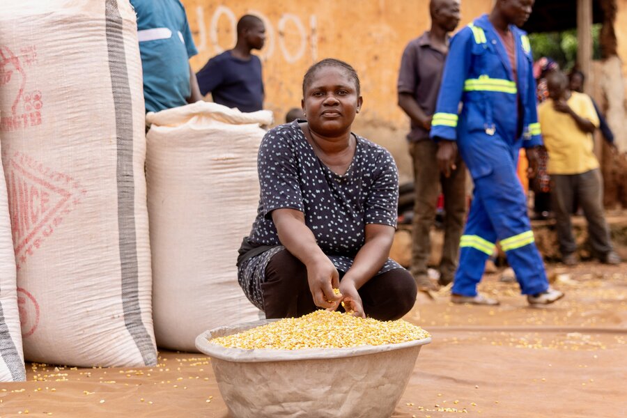 A woman squats next to a large metal bowl of maize, backdropped by feedbags and workers. Photo: Abdul-Wahab Mohammed
