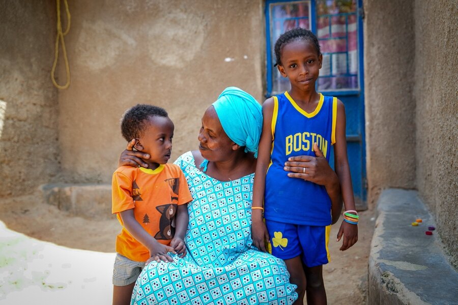 A woman in a blue dress and scarf sits in front of her house, clasping a child on either side of her. Photo: WFP/Daisy Masembe