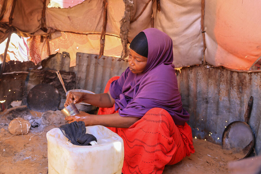 A woman in a red robe and purple headscarf sits in a tent, siphoning liquid from a plastic jug. Photo: WFP/CRC/Mohamed Ali