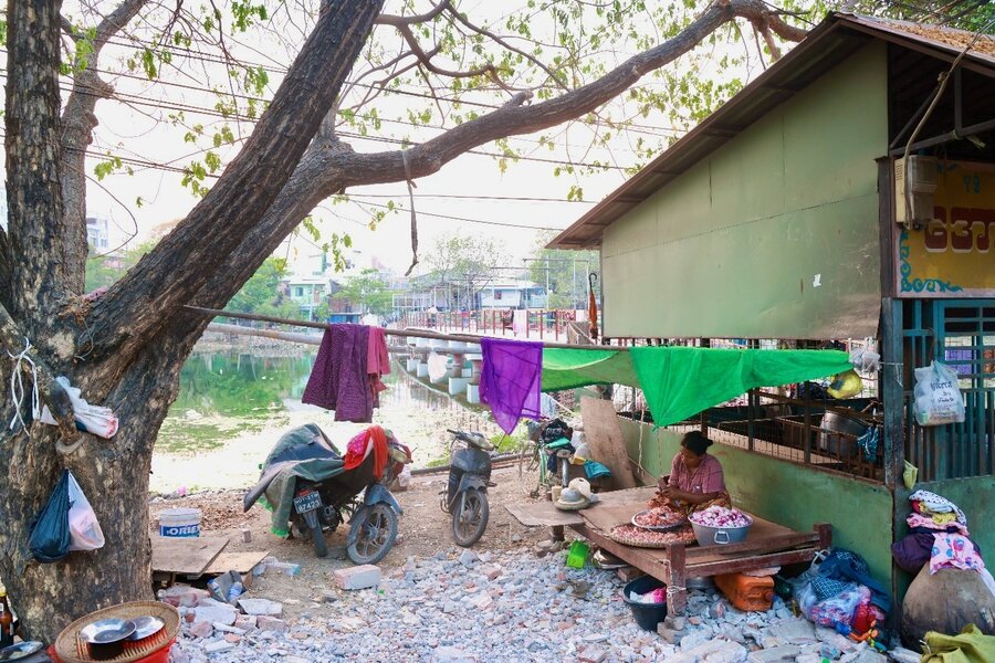 A woman chips vegetables outside, surrounded by motorcycles and clothes hanging from a line. Photo: WFP/Htet Oo Linn