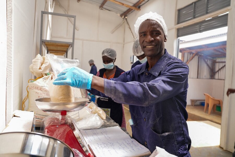A smiling man in a white cap and blue uniform weighs a plastic bag of formula on a scale. Photo: WFP/Daisy Masembe