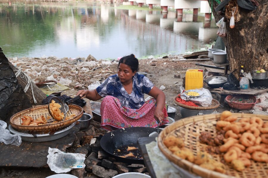 A woman in a long skirt and blue sits on the ground, stirring fritters she is cooking at her makeshift stall. Photo: WFP/Htet Oo Linn