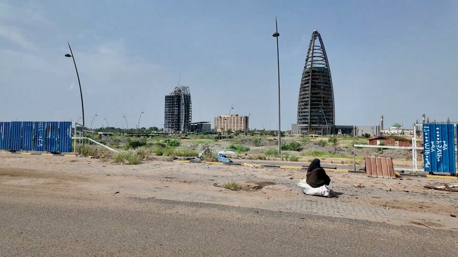 A woman sits next to a dusty road, backdropped by war shattered buildings. Photo: WFP/Leni Kinzli