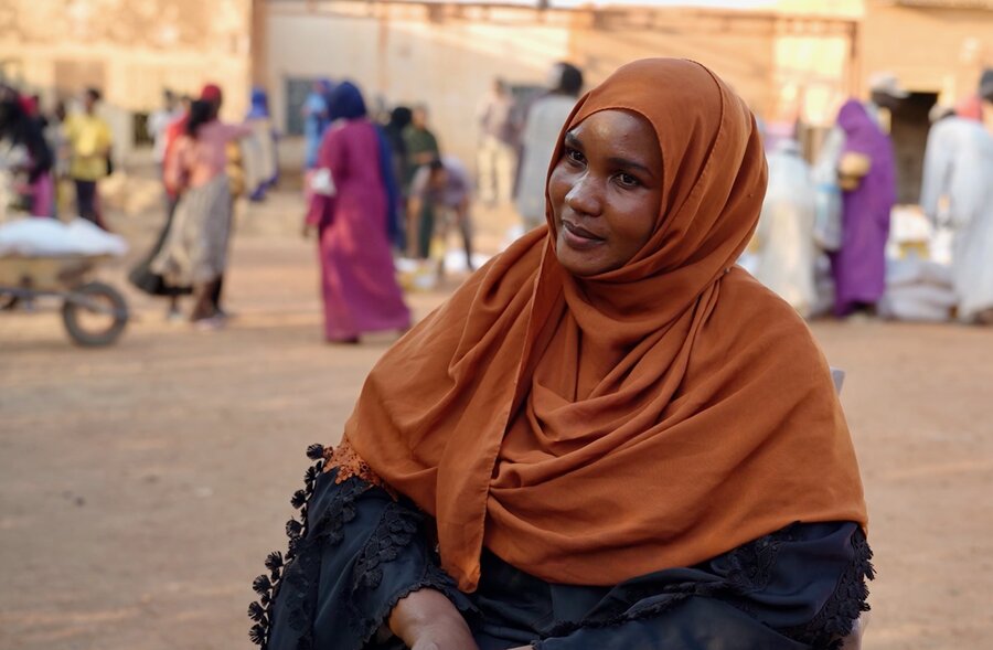 Closeup of a Sudanese woman in a brown headscarf and black robe backdropped by people in long robes walking around a dusty landscape. Photo: WFP