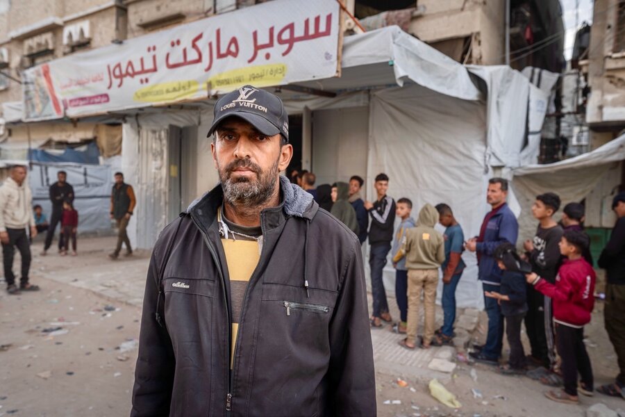 A man in a baseball cap and dark jacket stands in front of a shop partially covered in plastic, with a long line of customers. Photo: WFP/Maxime Le Lijour