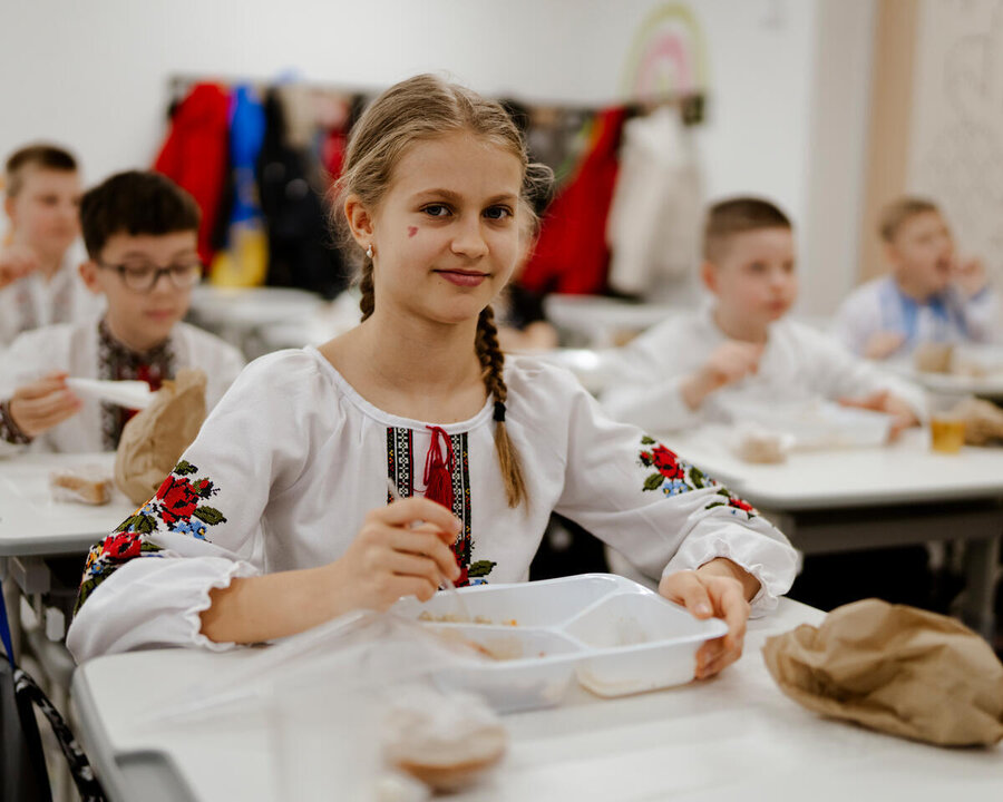 A child sits at a classroom table eating a meal from a divided tray, with other children eating at desks in the background.