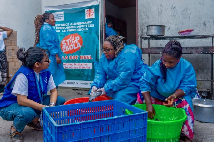 A woman wearing a blue WFP vest speak to two other women dressed in blue gowns preparing food. Behind them is a sign about the fight to stop Ebola. Photo: WFP/Benjamin Anguandia