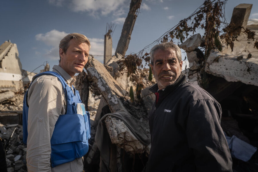Two people stand amid collapsed buildings and rubble one wearing a blue vest surveying destruction in a conflict-affected urban area under cloudy sky.