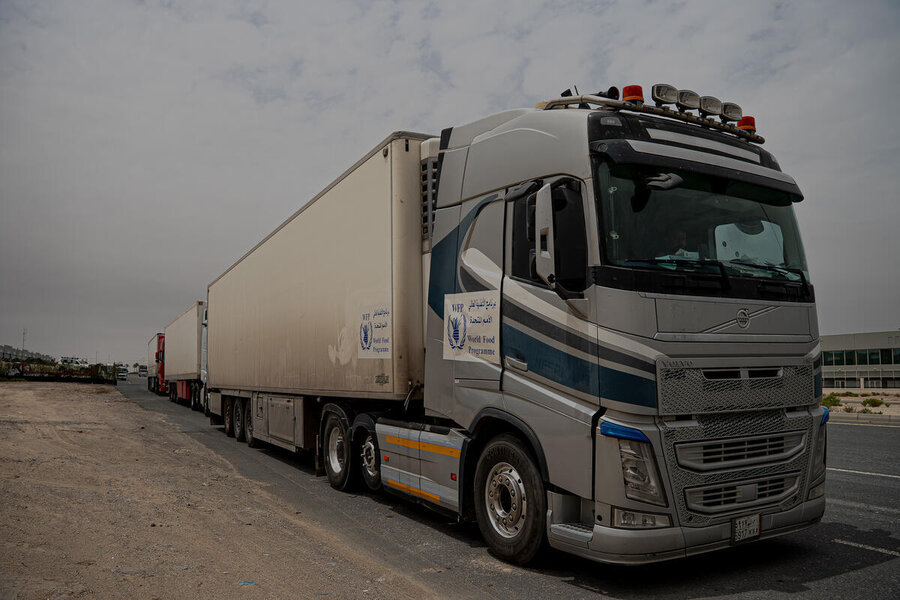WFP-branded truck with long trailer parked roadside, part of a convoy transporting humanitarian food supplies along a highway in an arid area.