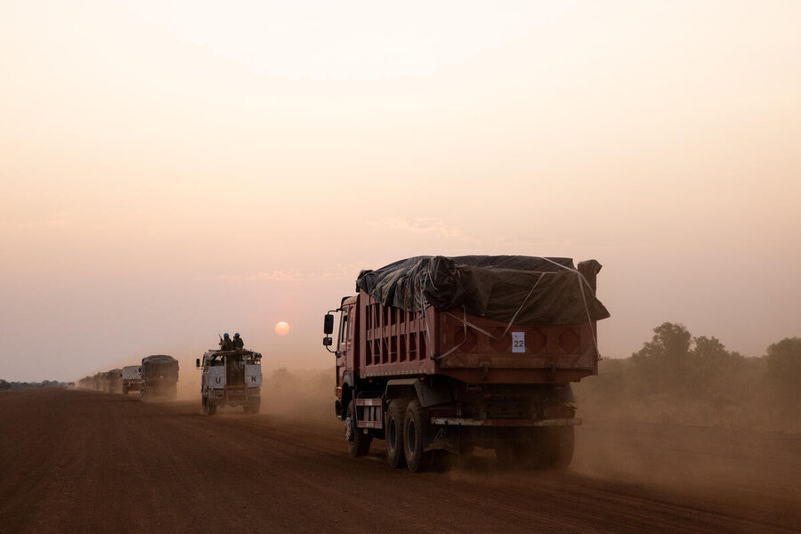 Humanitarian aid convoy of trucks travels along a dusty road at sunset, raising clouds of dust as vehicles move supplies across a remote landscape.