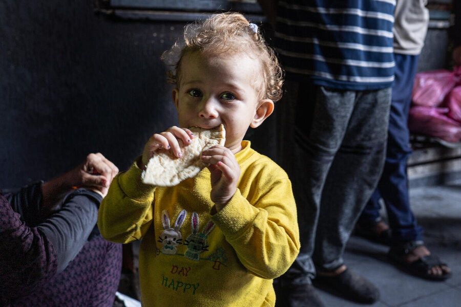 Copilot said:  A young child in a yellow sweater eats flatbread indoors while other people stand nearby, illuminated by soft natural light in a dim room.