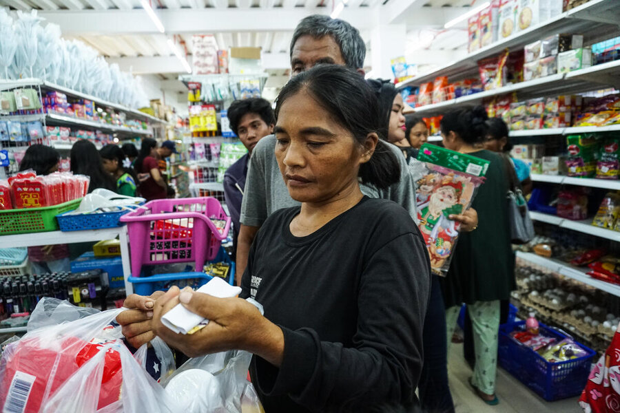 People shop in a busy grocery store, selecting essential items from shelves and checking purchases at the checkout area amid a crowded aisle.