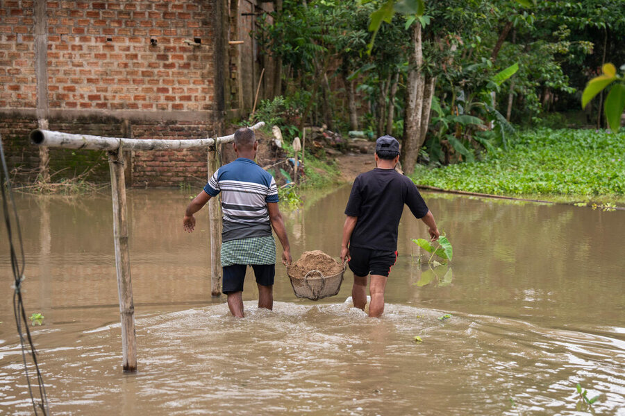 Copilot said:  Two people wade through floodwater carrying a basket of soil, moving past submerged homes and vegetation in a waterlogged community.