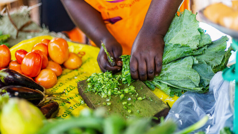 Hands chop leafy green vegetables on a wooden board beside tomatoes and eggplants, preparing fresh ingredients for a nutritious meal.