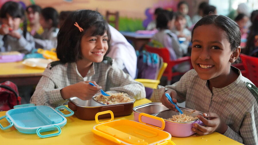 Two schoolchildren sit at a classroom table eating lunch from reusable containers, while other pupils gather in the background of a brightly coloured room.