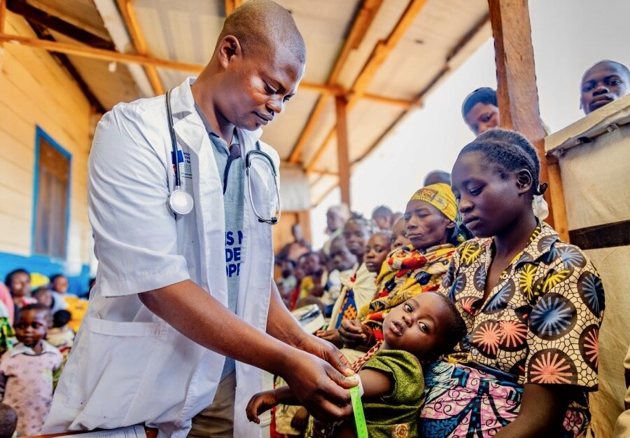 A male nurse with a stethoscope around his neck, checks a baby for malnutrition in a crowded health centre. Photo: WFP/Charly Kasareka