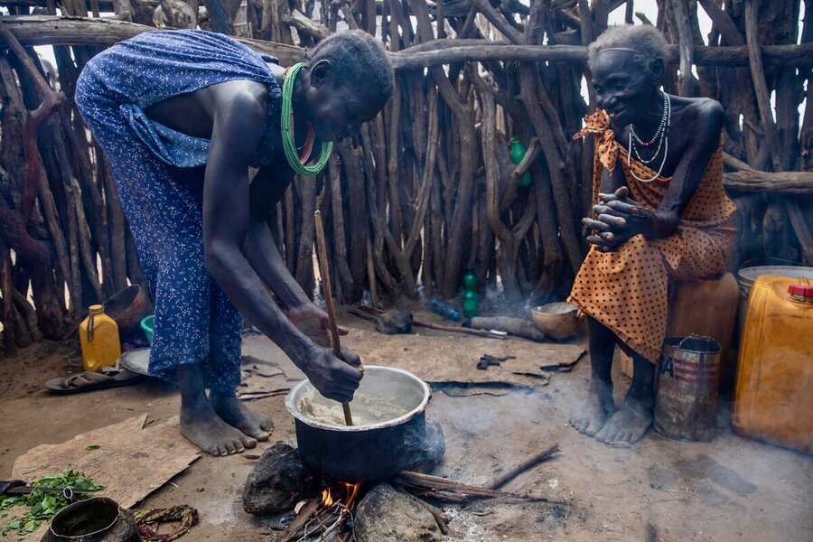 A women bends over an open fire stirring a porridge with a stick, while another sits nearby on a plastic jug watching her and smiling. Photo: WFP/Gabriela Vivacqua