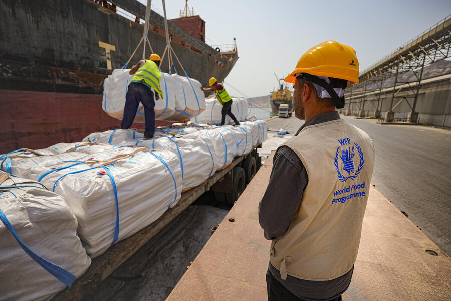 WFP staff supervise the transfer of large aid cargo bags from a ship to trucks at a port loading facility